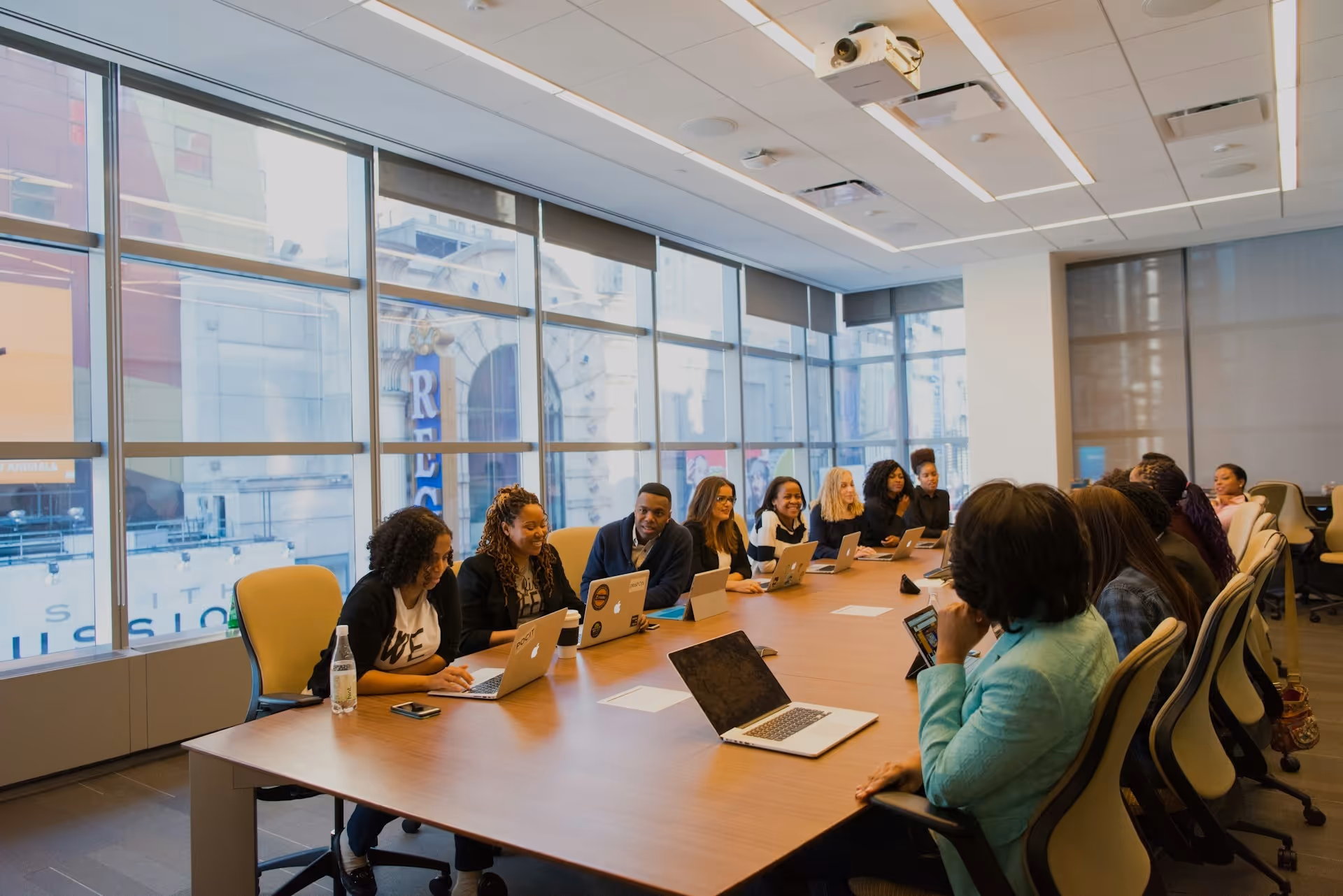 Group of people having a meeting around a long table with laptops in a modern conference room with large windows.