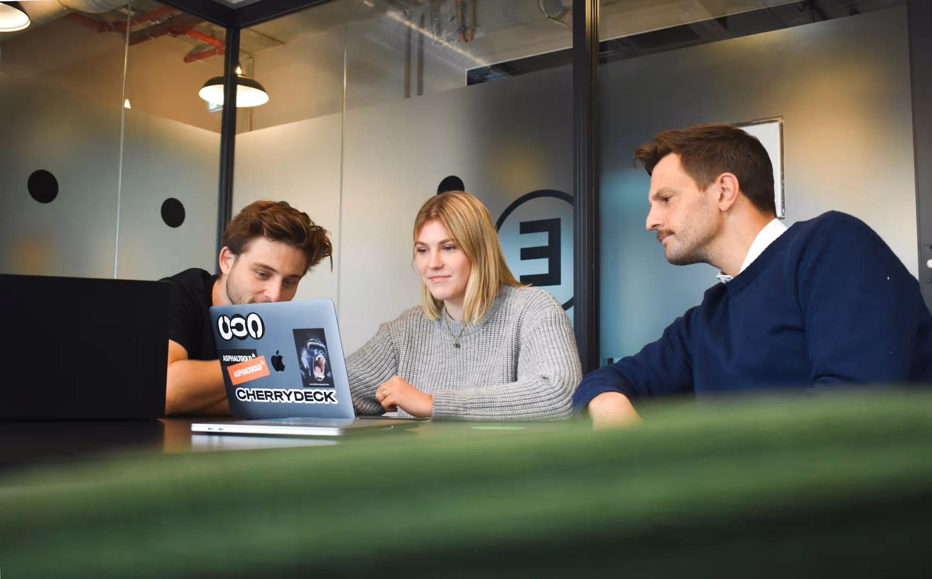 Three young professionals collaborating while looking at a laptop with stickers in a modern office.