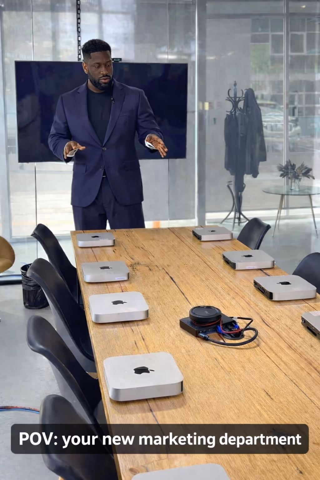 Man in a navy suit standing at the end of a conference table with several Mac Mini computers arranged on it in a modern office room.