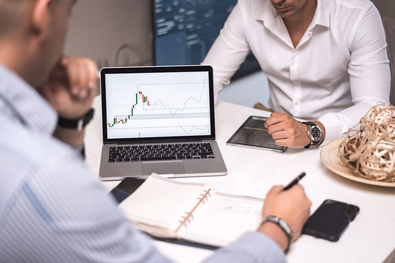 Two men in business attire reviewing stock market charts displayed on a laptop and tablet at a white desk with notebooks and a decorative basket.