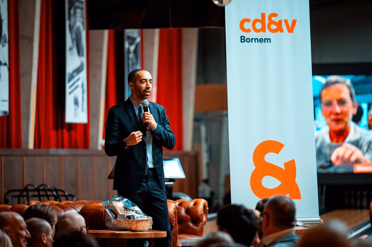 Man in a suit speaking into a microphone at an event with a cd&v Bornem banner and an audience.
