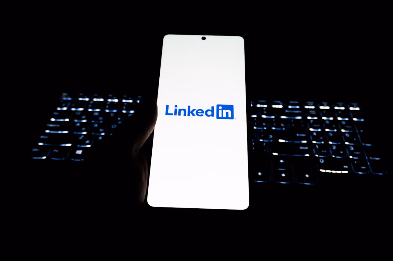 Hand holding smartphone displaying the LinkedIn logo in front of a backlit keyboard in darkness.
