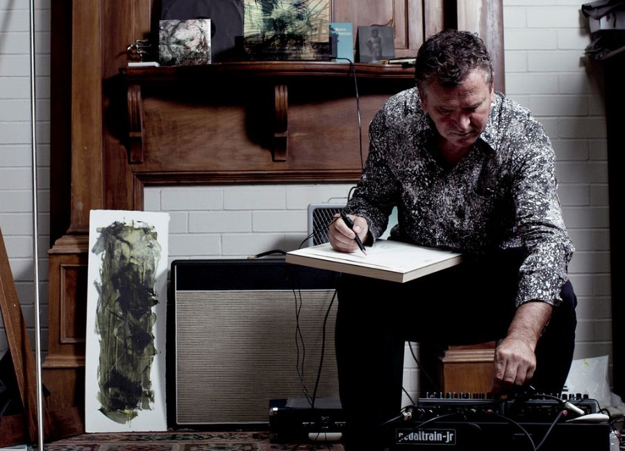 Dr Jon Tarry seated in his studio, holding a sketchpad and pen, adjusting audio equipment with abstract paintings visible around him.