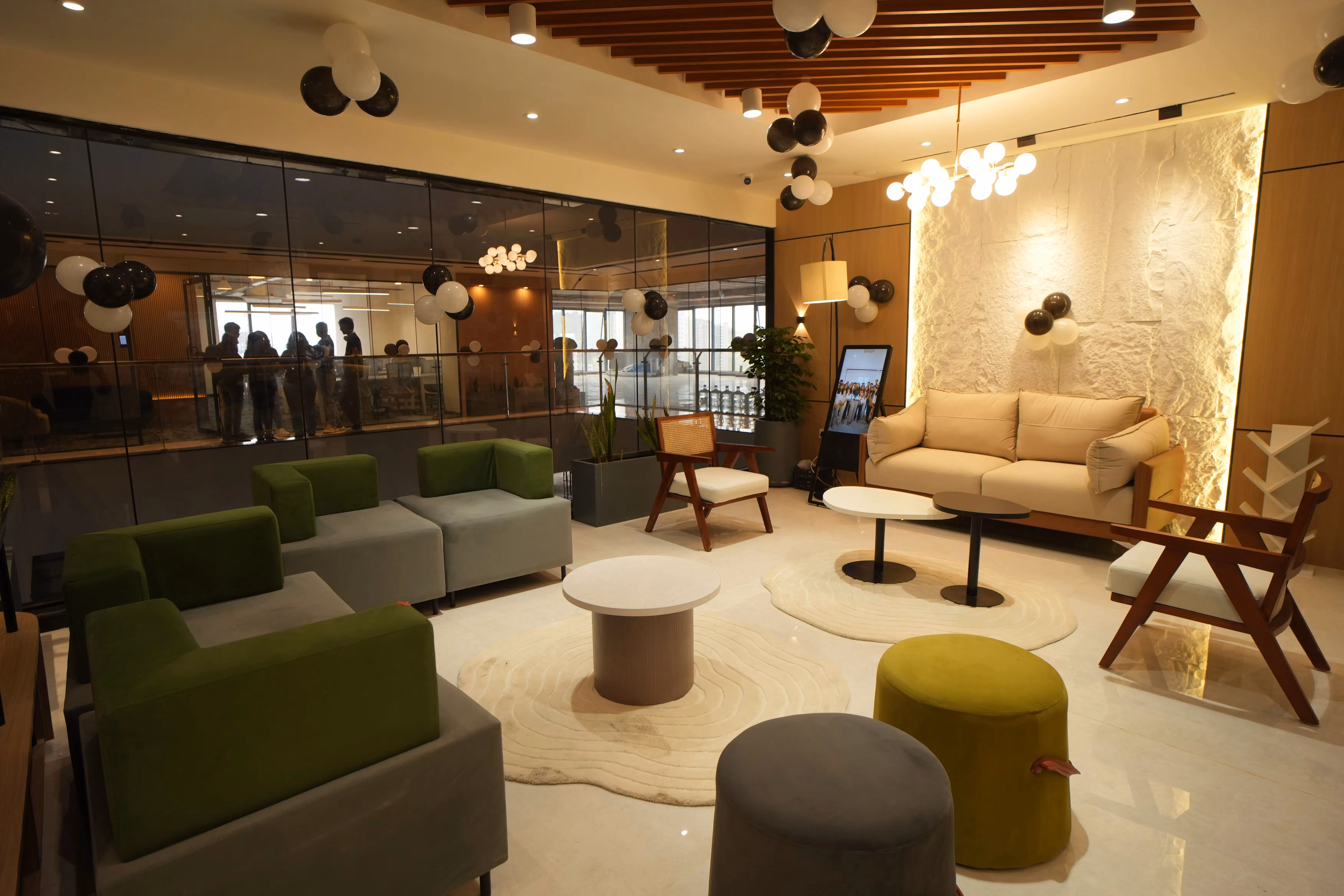 Modern lounge area with green and beige sofas, wooden chairs, round tables, black and white balloons, and a large glass wall showing people in an adjacent room.