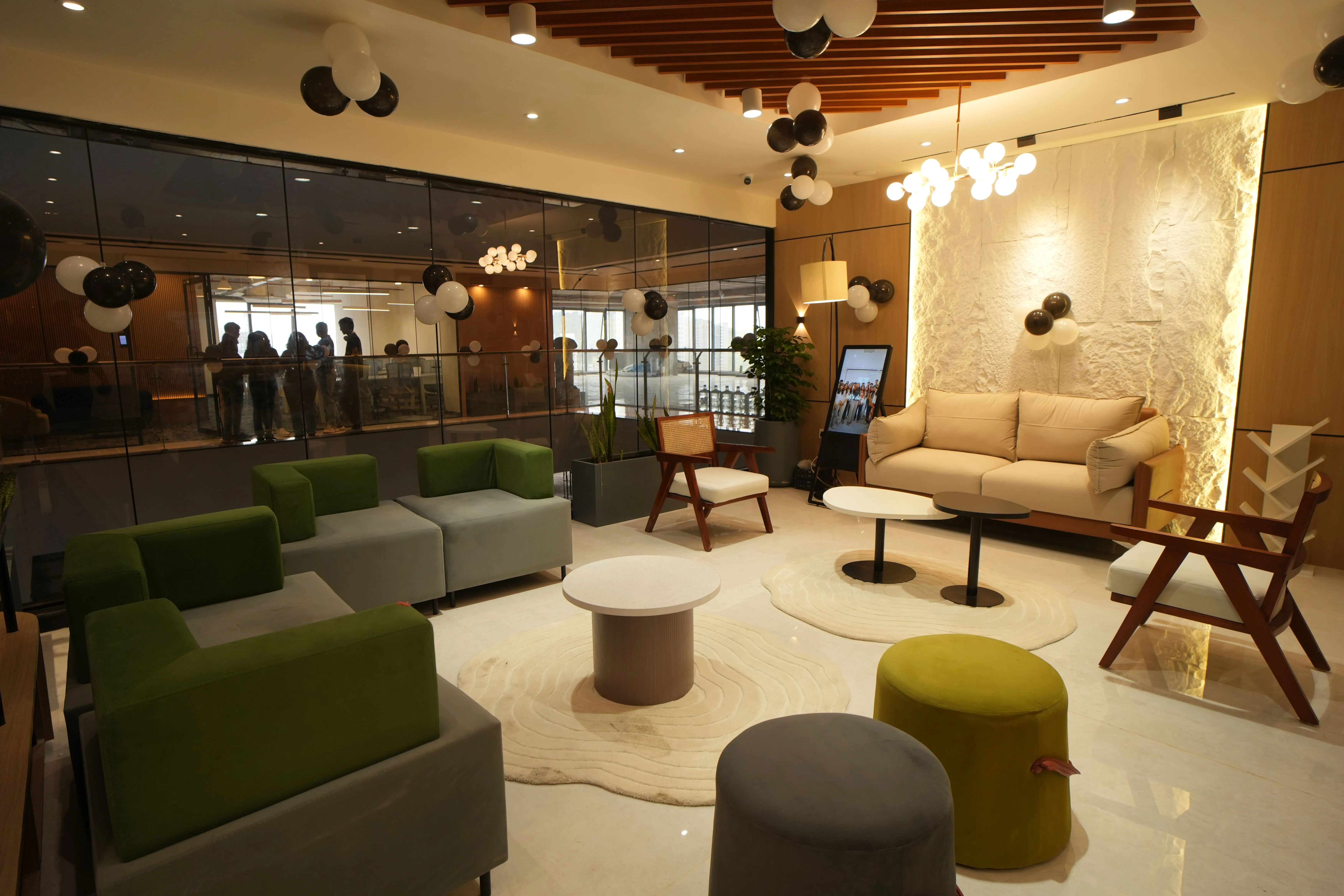 Modern lounge area with green and beige sofas, wooden chairs, round tables, black and white balloons, and a large glass wall showing people in an adjacent room.