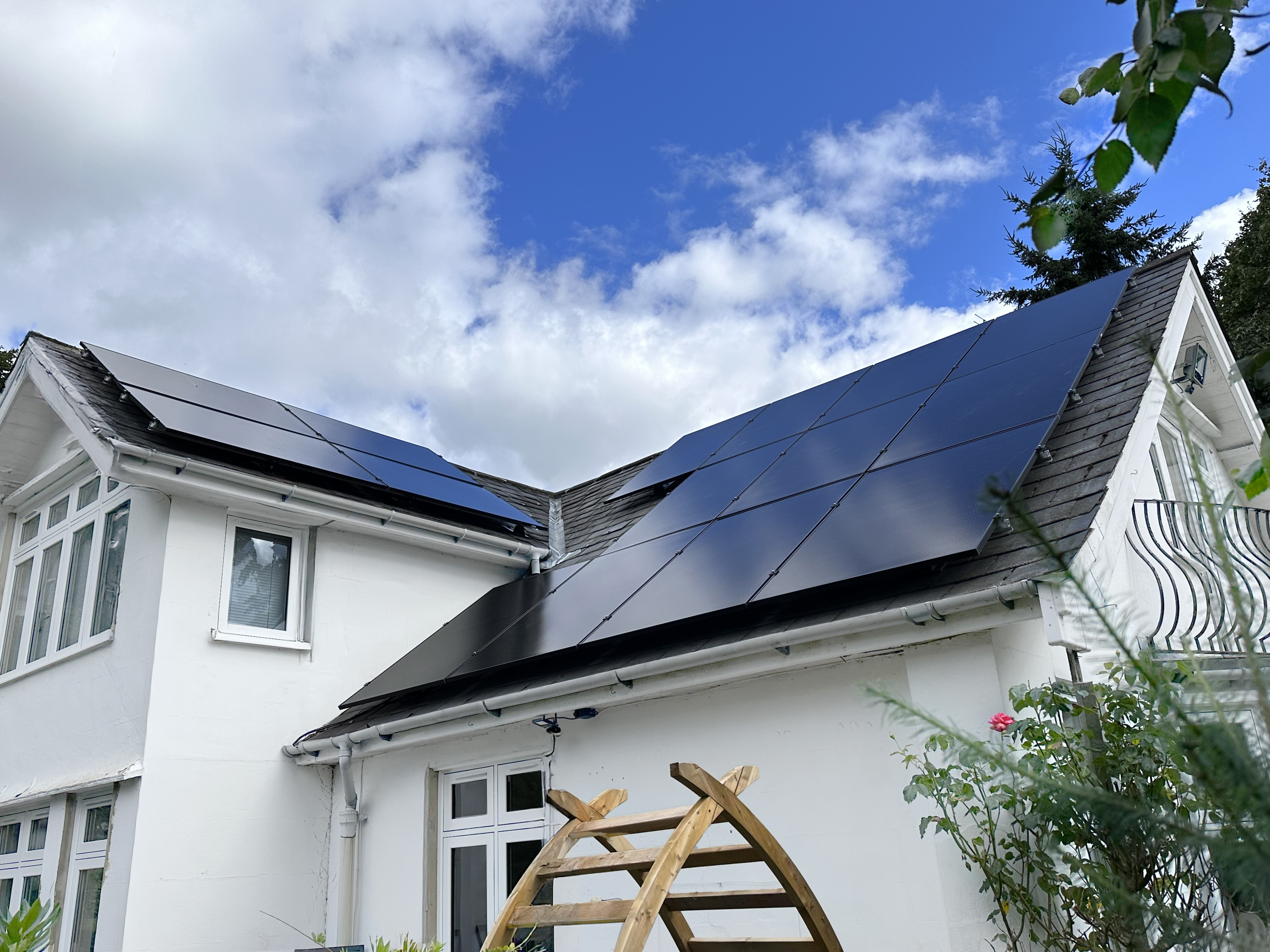 White house with multiple solar panels installed on the sloped roof under a partly cloudy blue sky.