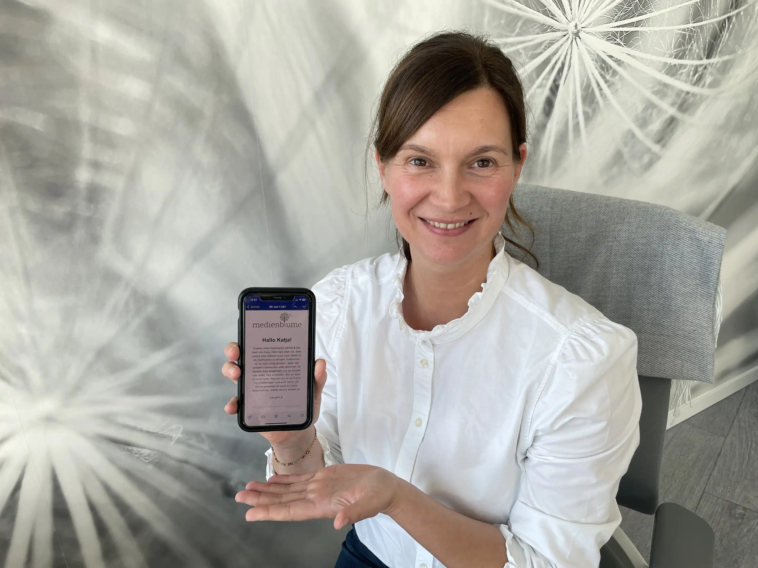 Frau in weißer Bluse sitzt auf Stuhl und zeigt lächelnd ein Smartphone mit einer medienblume E-Mail.