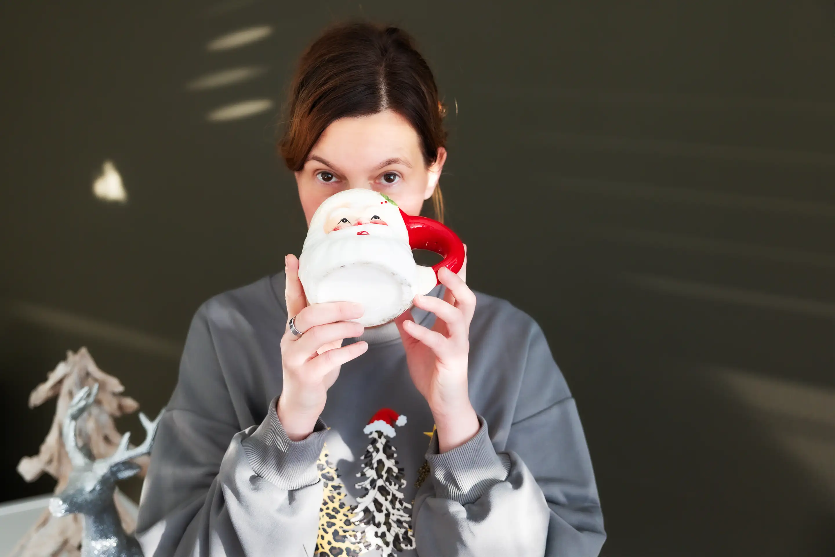 Frau in grauem Pullover hält eine Tasse mit Weihnachtsmann-Motiv vor ihr Gesicht.
