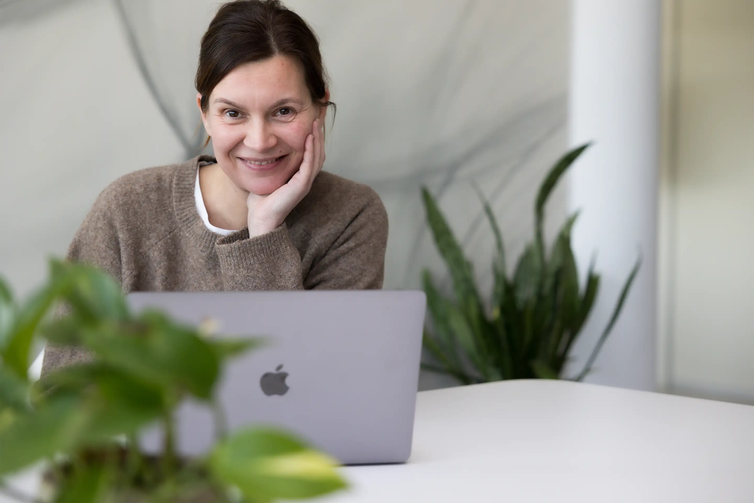 Lächelnde Frau in braunem Pullover sitzt an einem weißen Tisch mit offenem MacBook, Pflanzen im Vorder- und Hintergrund.