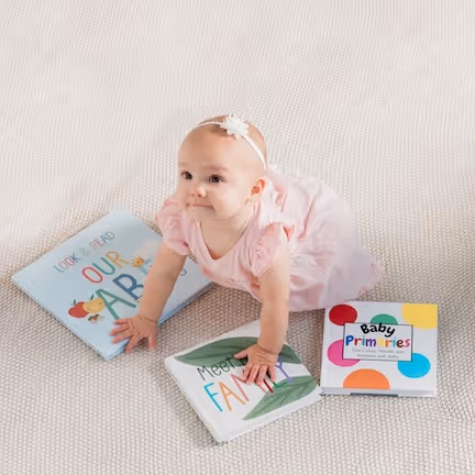 Baby girl in pink dress and headband crawling on carpet with three colorful Stareworthy children's books around her.