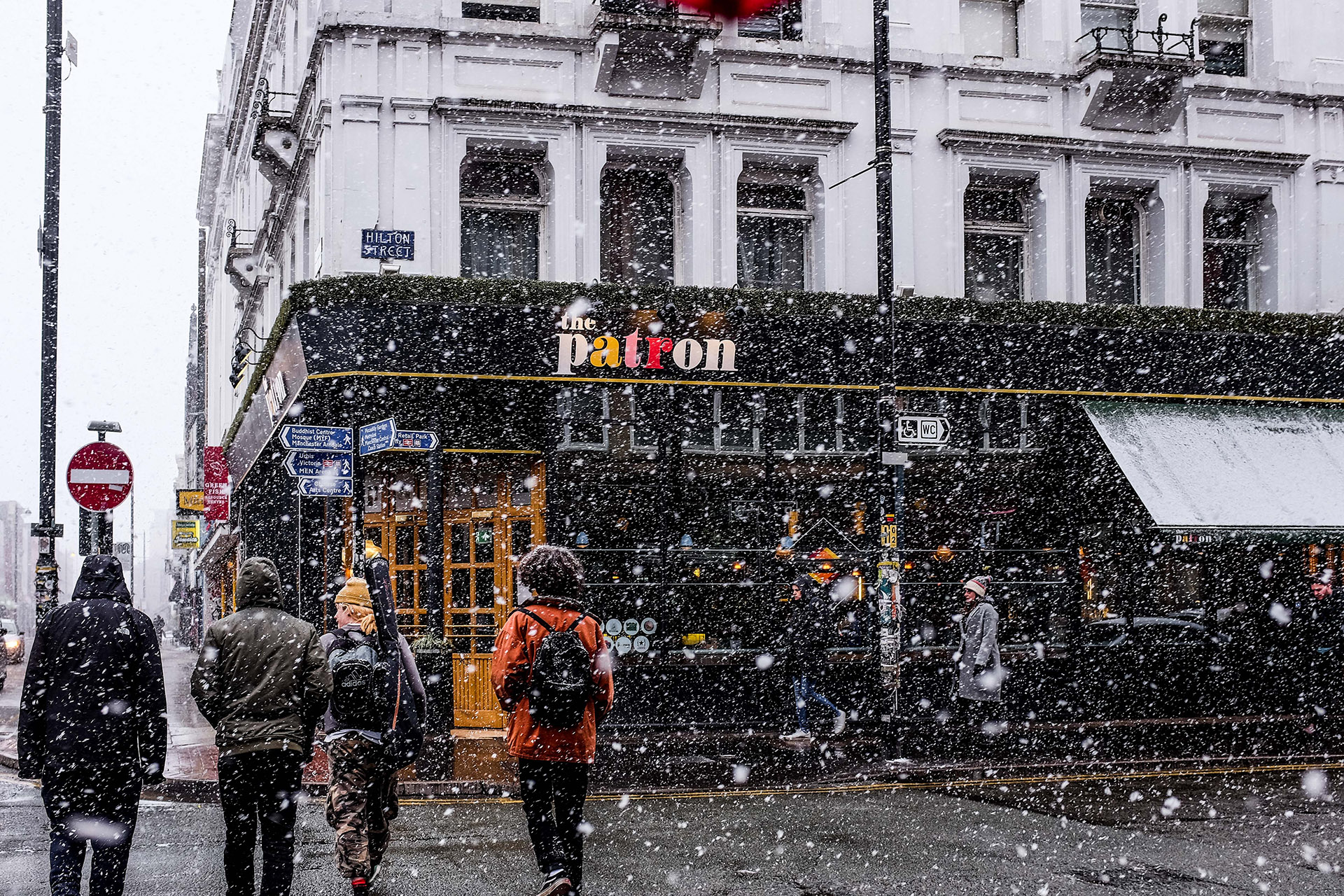 People crossing a snowy street with their backs to the camera; a building with a sign reading 'The Patron' is in the background.