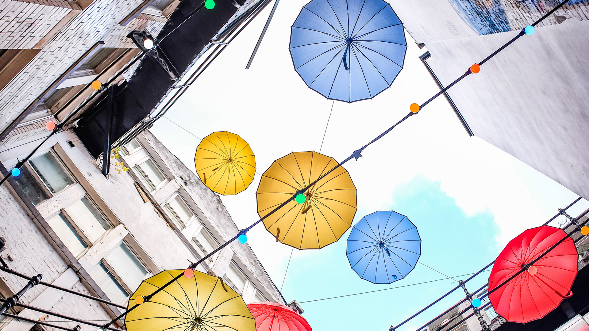 Red, blue, and yellow umbrellas hung in a courtyard, seen from below against a cloudy blue sky.