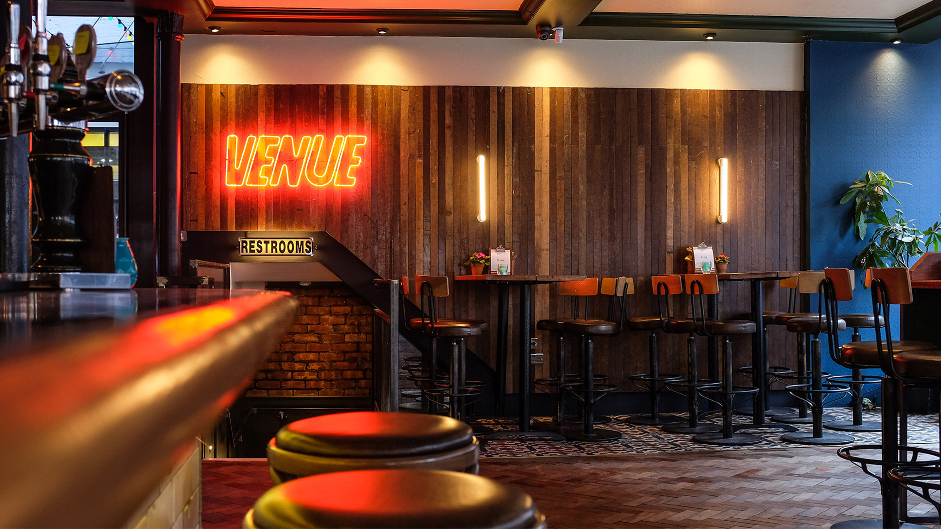 A neon sign reading 'Venue' on a wood-panelled wall, with two bar stools in the foreground.