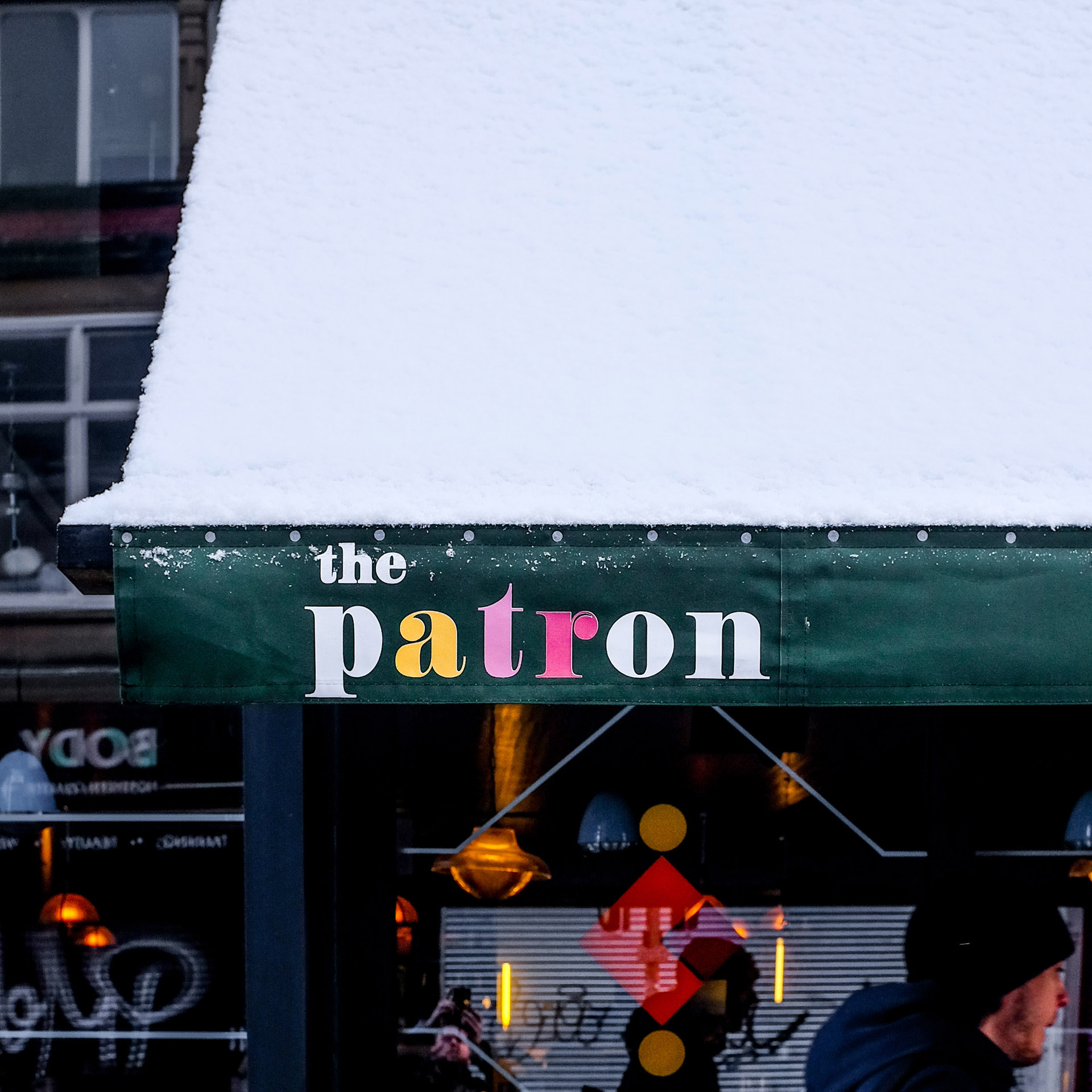 Close-up of a green awning with snow on top, featuring The Patron logo, with the Patron window graphics visible in the background.
