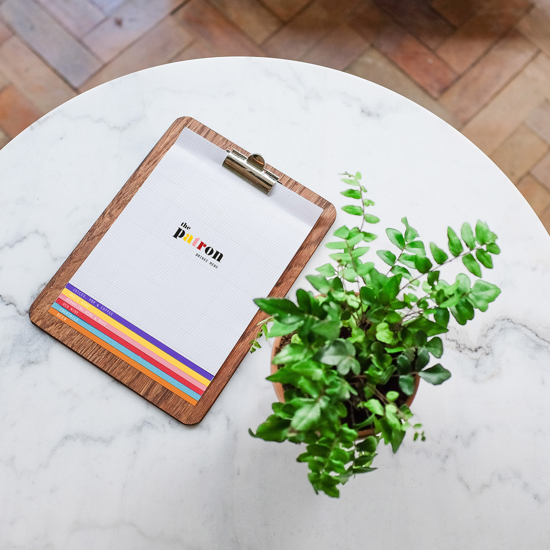 The Patron drinks menu on a wooden clipboard on a round marble table next to a potted plant.