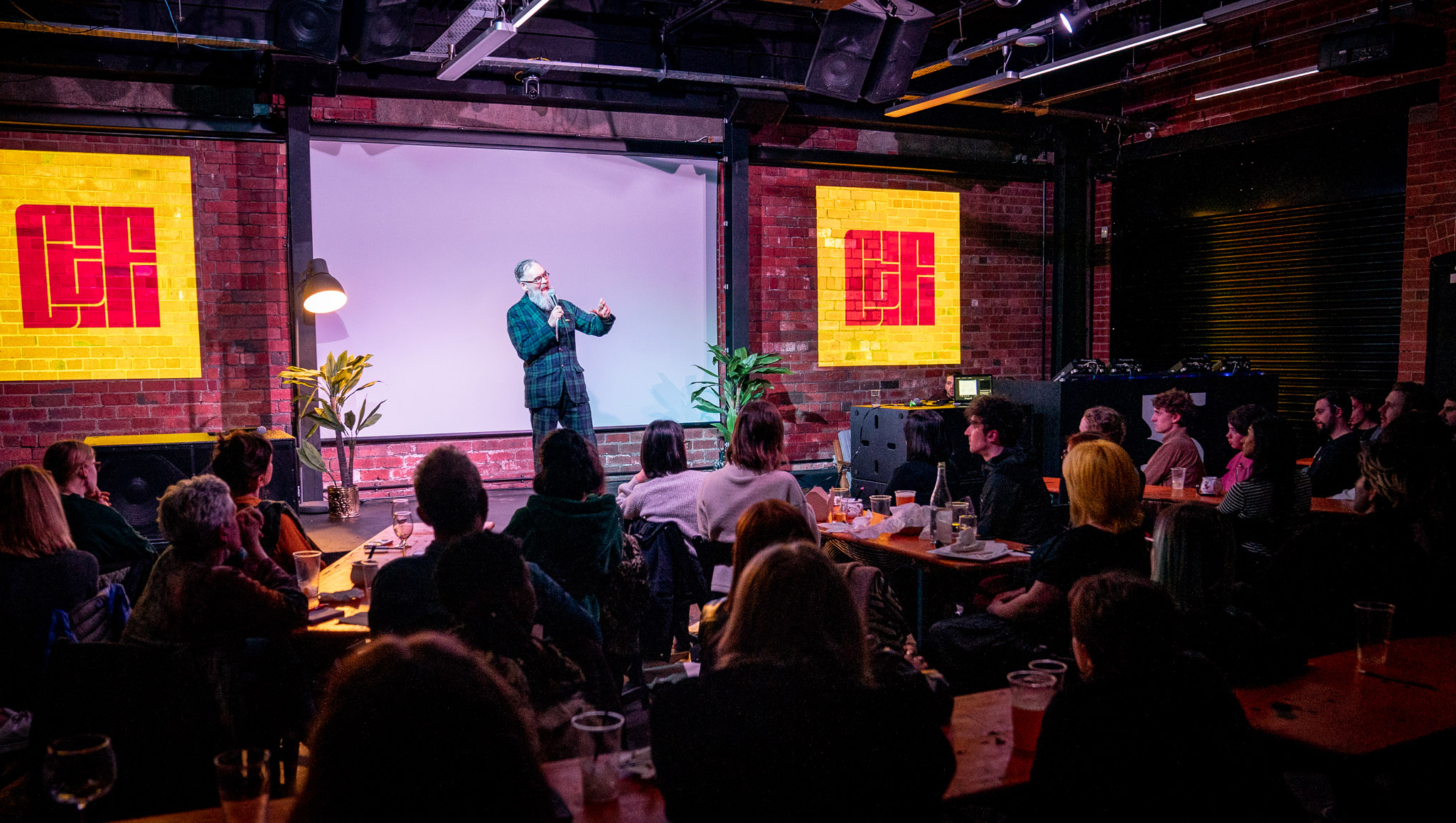 Older bearded man in a tartan suit speaks on stage, with the 'Chew the Fat' logo projected on a brick wall.