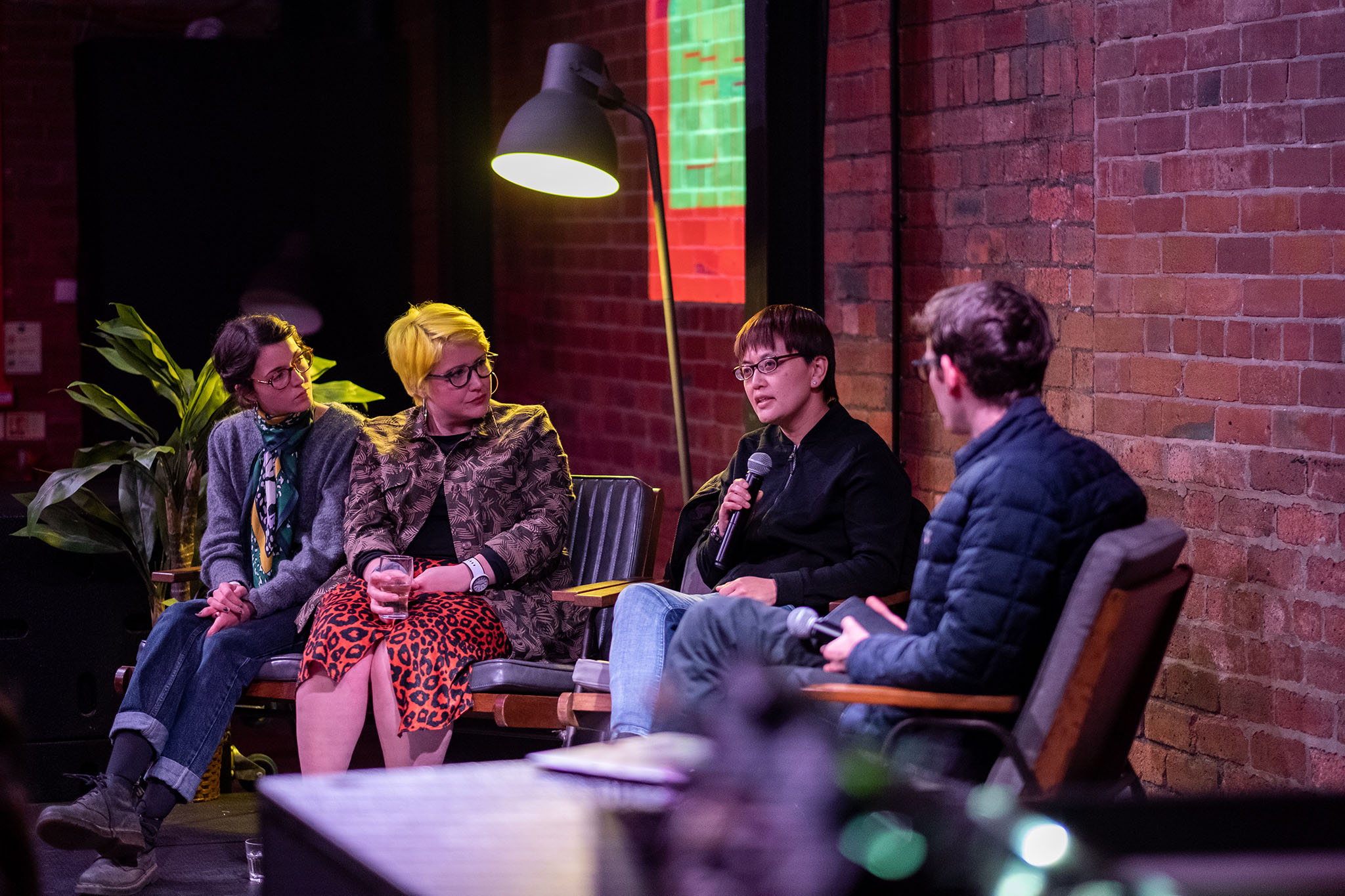 A panel of three women and one man sits on stage; three listen while one woman speaks into a microphone.