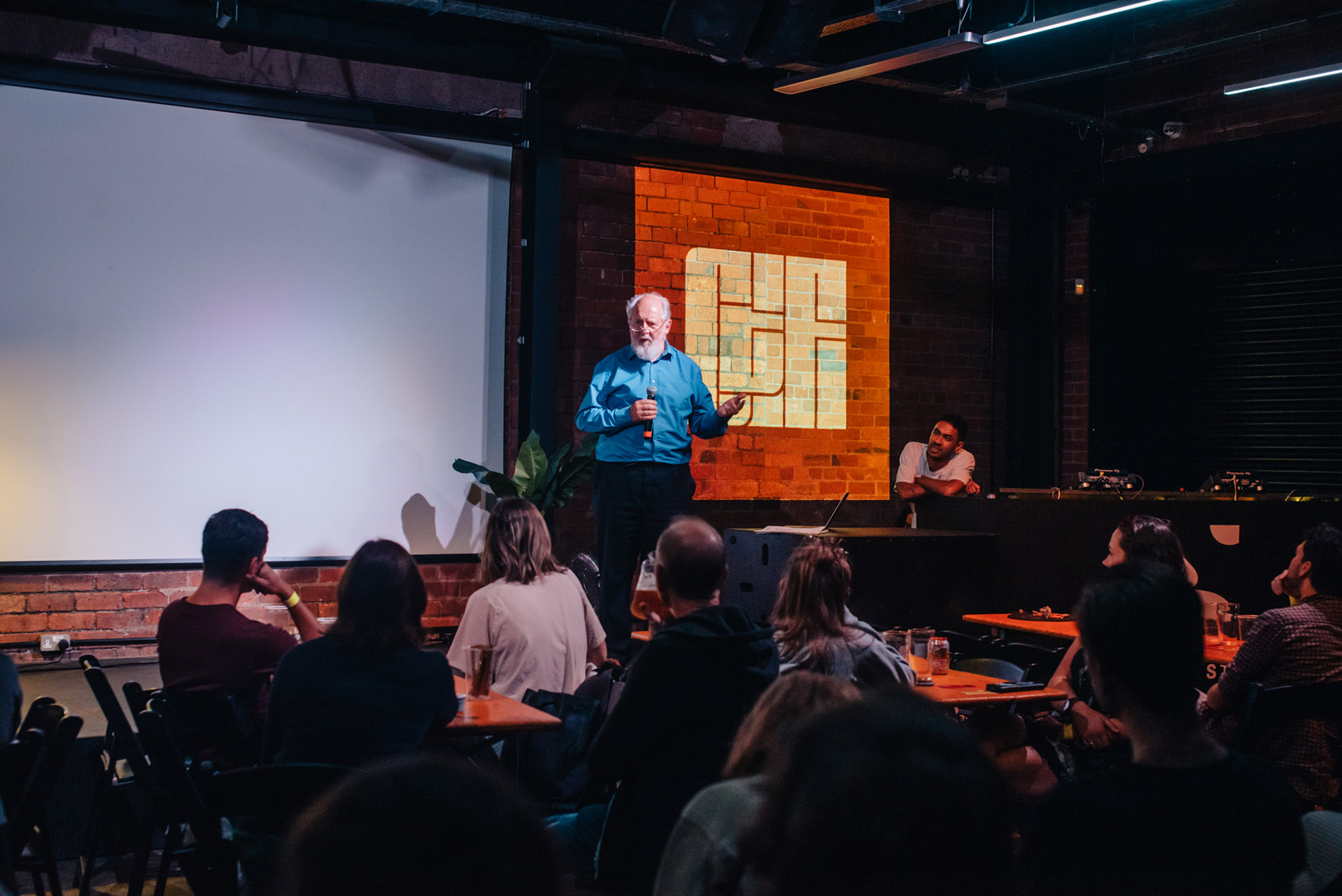 Audience watches an elderly man with a white beard and blue shirt presenting on stage.