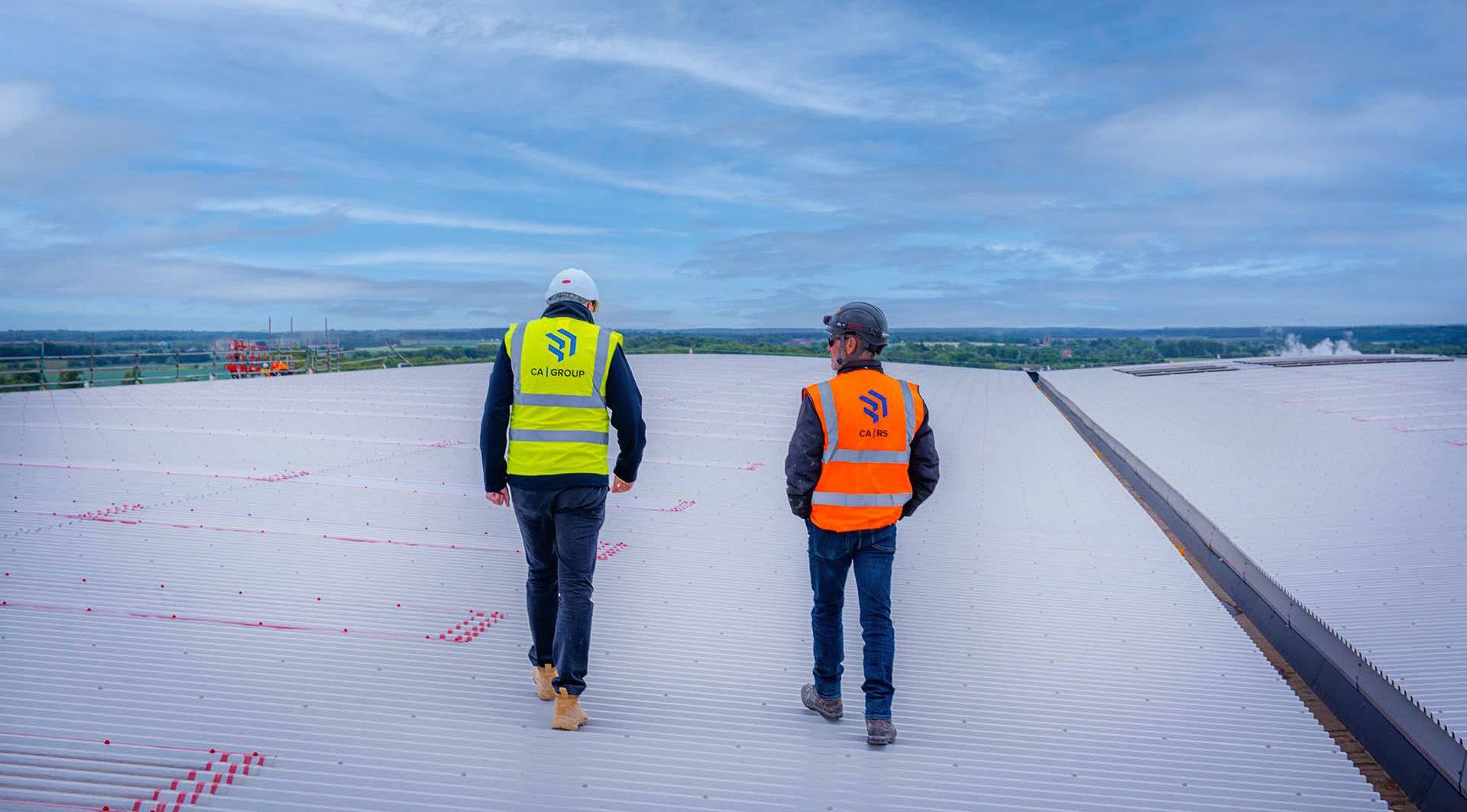 Two men wearing high-visibility jackets walking across the roof of a large building.