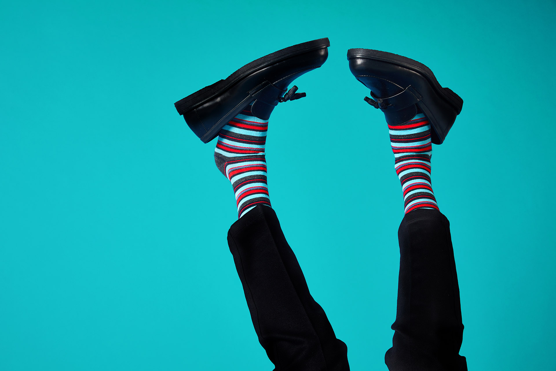 Legs raised in the air, wearing striped socks and black loafers against a teal backdrop.