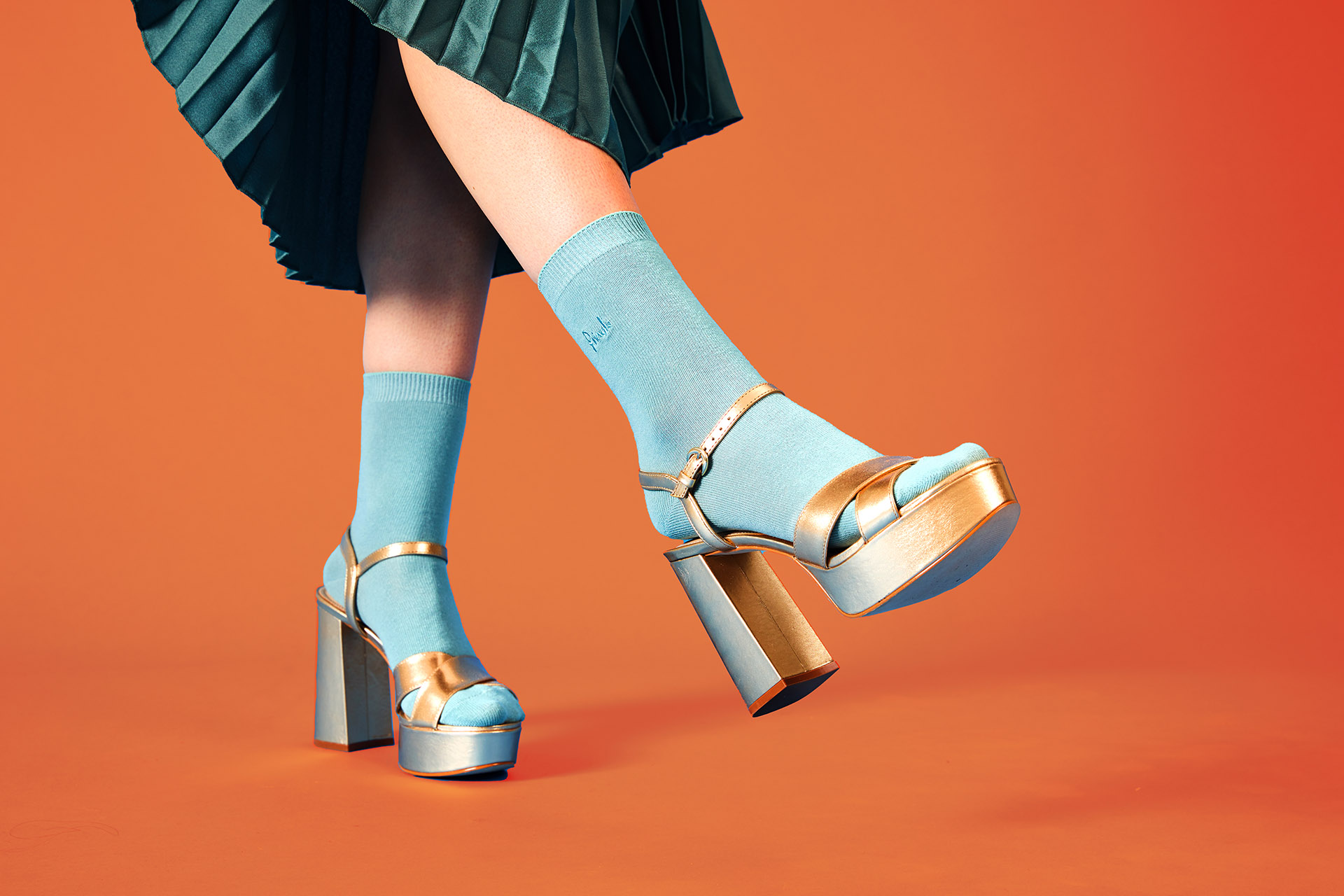 Close-up of a woman's lower body wearing gold heels, blue socks and a green skirt, kicking one foot into the air.