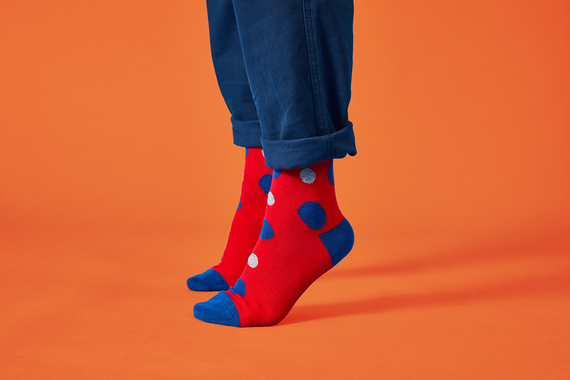 Close-up of legs wearing navy trousers and red socks with blue and white polka dots on an orange backdrop.