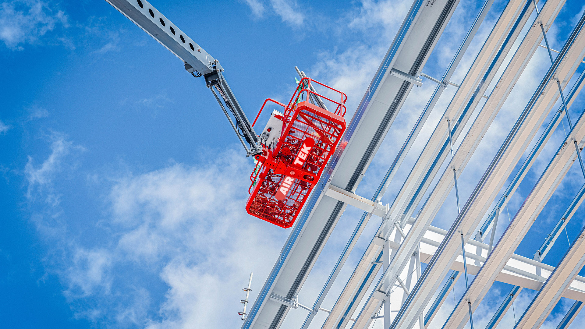 Looking up, a MEWP bearing the Airtek logo operates on a building against a cloudy blue sky.