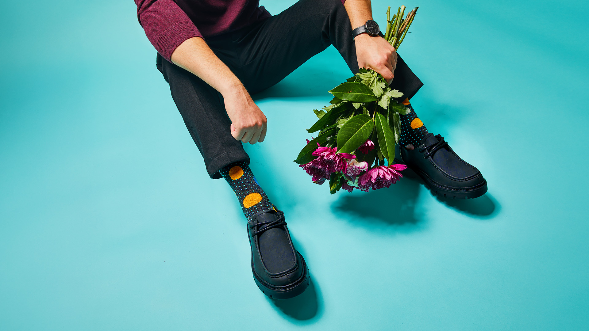 Close-up of a man sitting on the floor wearing black trousers and polka-dot socks, holding flowers on a turquoise background.