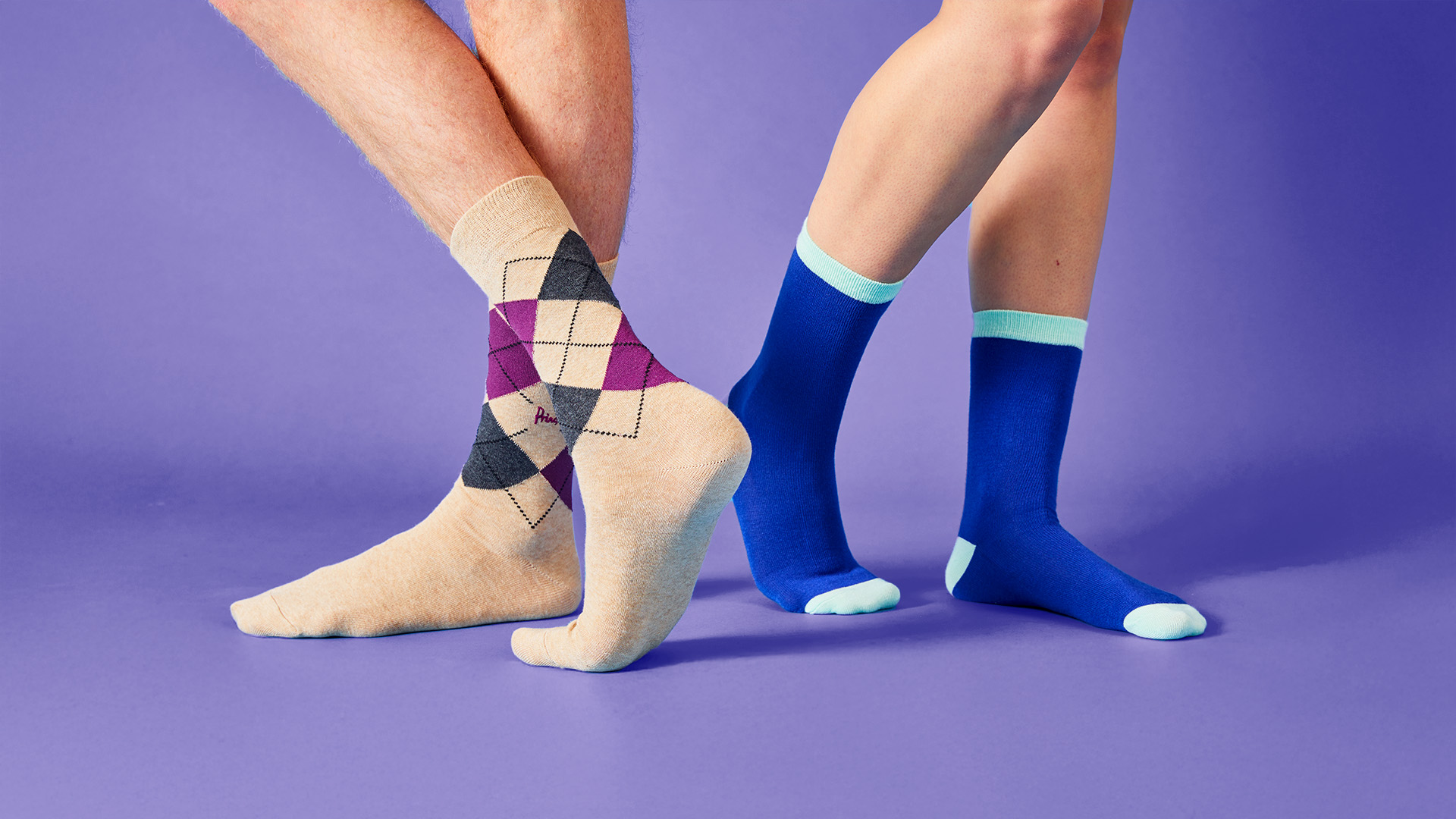 Close-up of two people’s legs on a lilac backdrop: one wearing dark blue socks, the other beige socks with a diamond pattern.