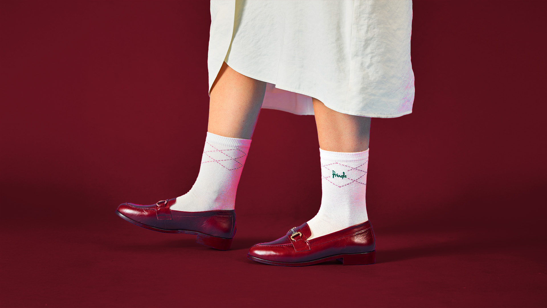 Close-up of a woman wearing white Pringle socks with pink lines, burgundy loafers and a white long skirt on a burgundy background.