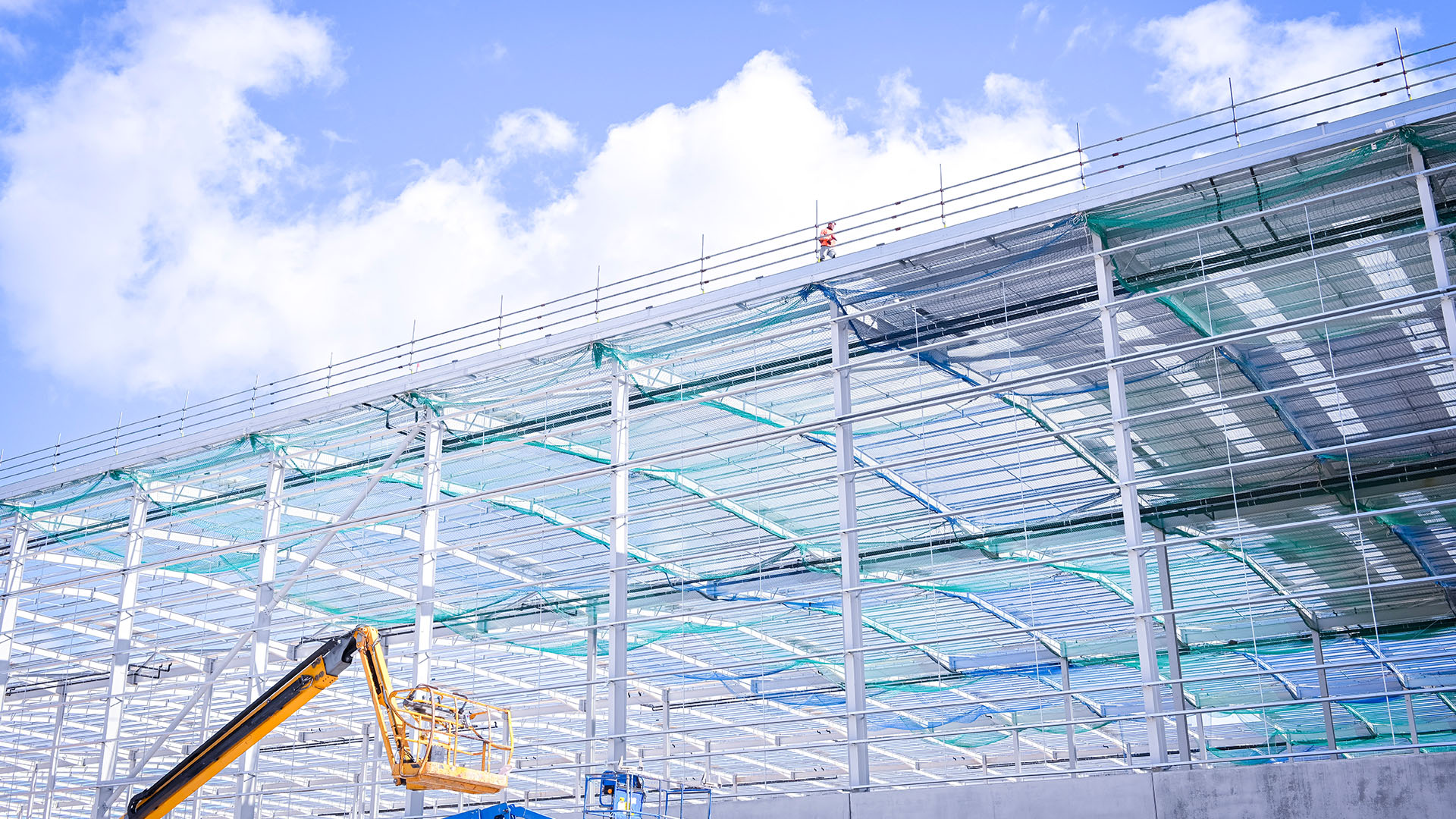 Industrial building under construction with a boom lift in front and a man on the roof in a high-visibility jacket.