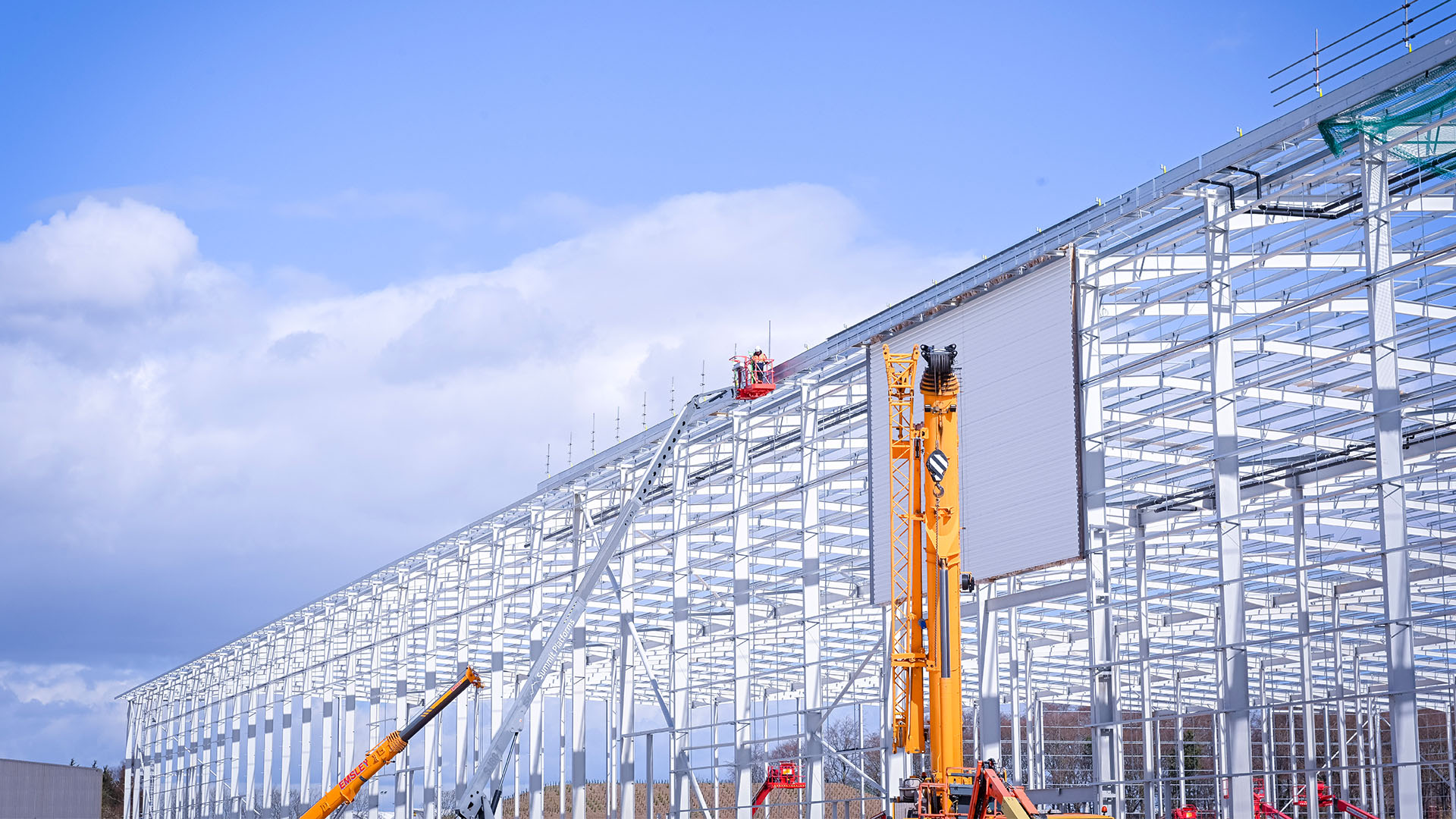 Man on a boom lift at the side of an industrial building under construction, with a cloudy blue sky in the background.