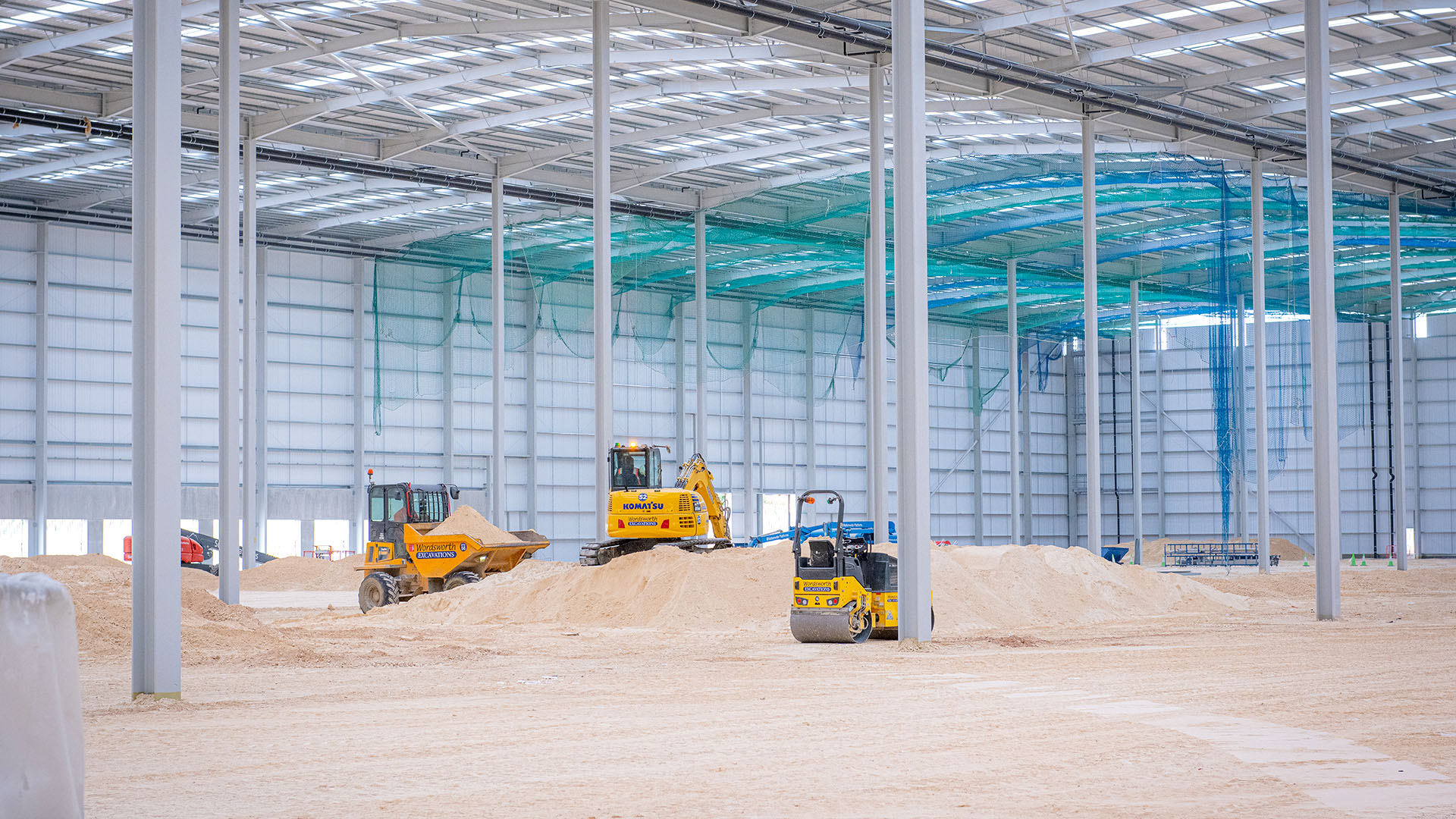 Three yellow construction vehicles on a pile of dirt inside an industrial building under construction, with safety netting in the background being removed.
