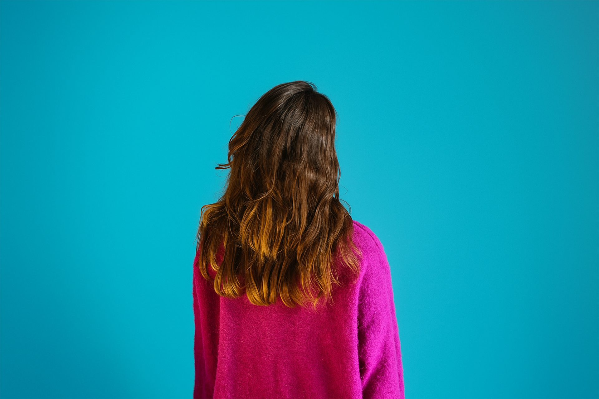 A woman wearing a purple jumper with her back to the camera on a teal backdrop.