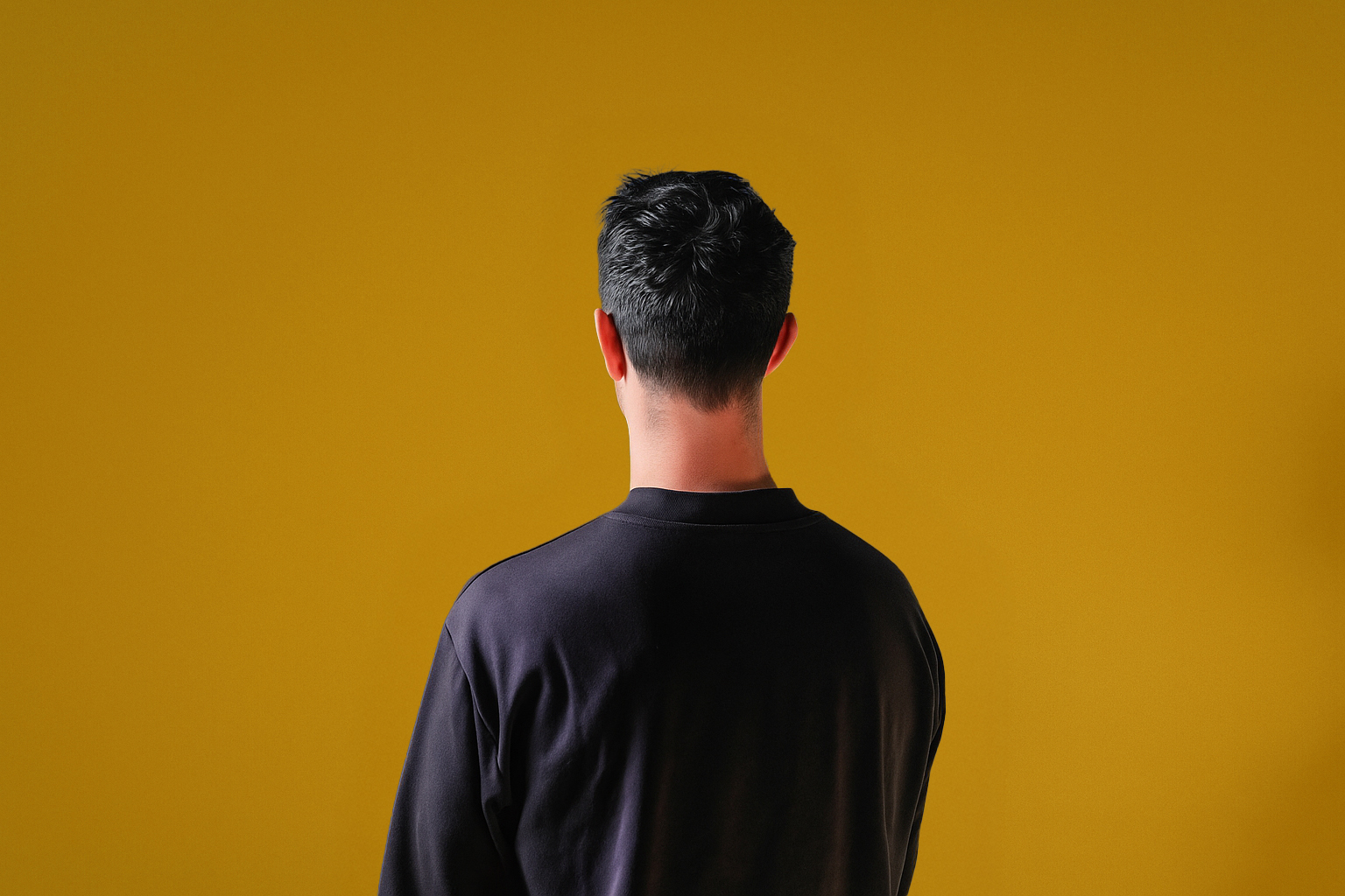 A man wearing a black t-shirt with his back to the camera on a dark yellow backdrop.