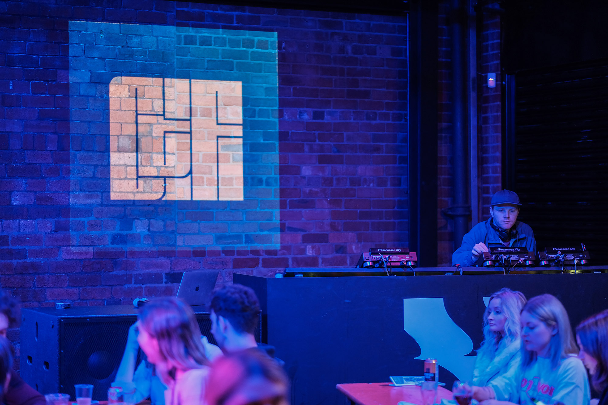 People sitting in the foreground while a man wearing a cap DJs in the background.