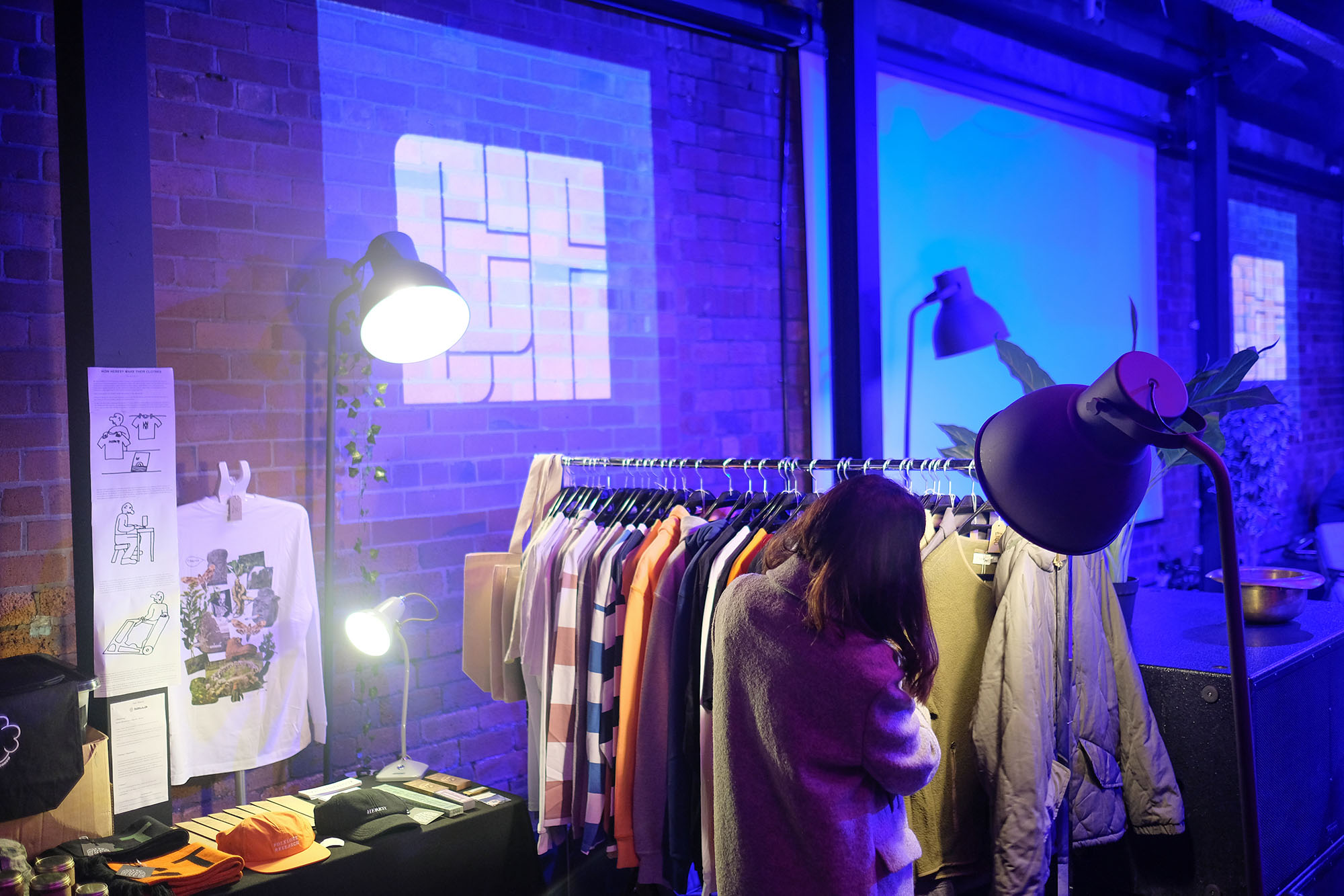 A woman browses a stall selling clothes and merchandise, with the Chew the Fat logo projected onto a brick wall in the background.