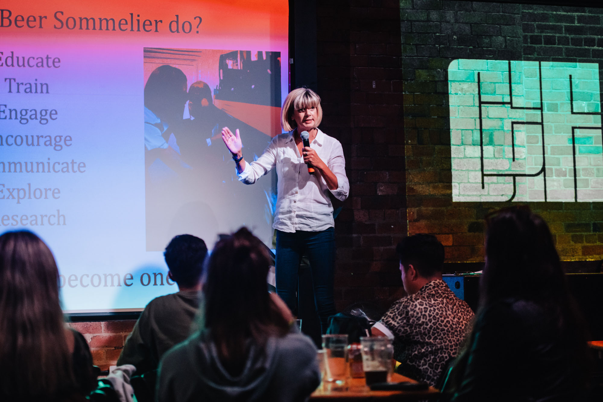 Audience watches as a woman speaks into a microphone on stage in front of a projected presentation.