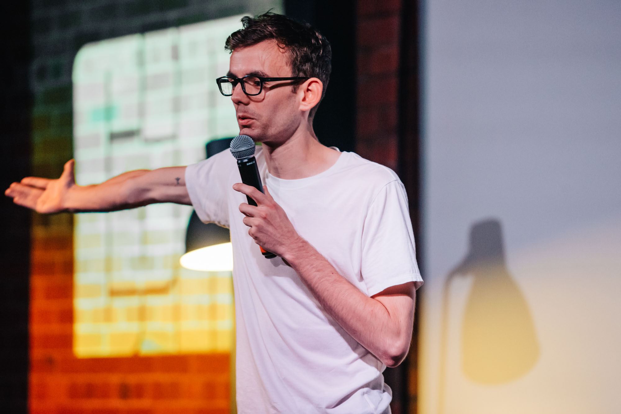 Close-up of a man wearing glasses speaking into a microphone on stage, with his arm outstretched.