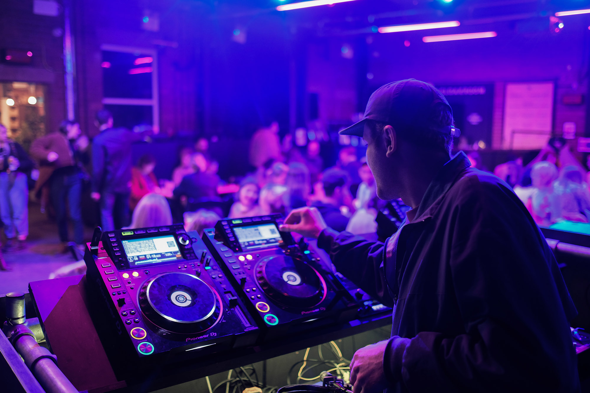 Man DJ-ing, wearing a cap, with his back to the camera, while people sit in the background.