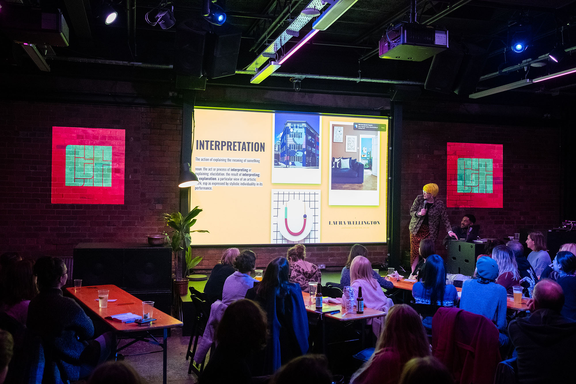 A woman stands to the right of the stage as she speaks in front of a projected presentation.