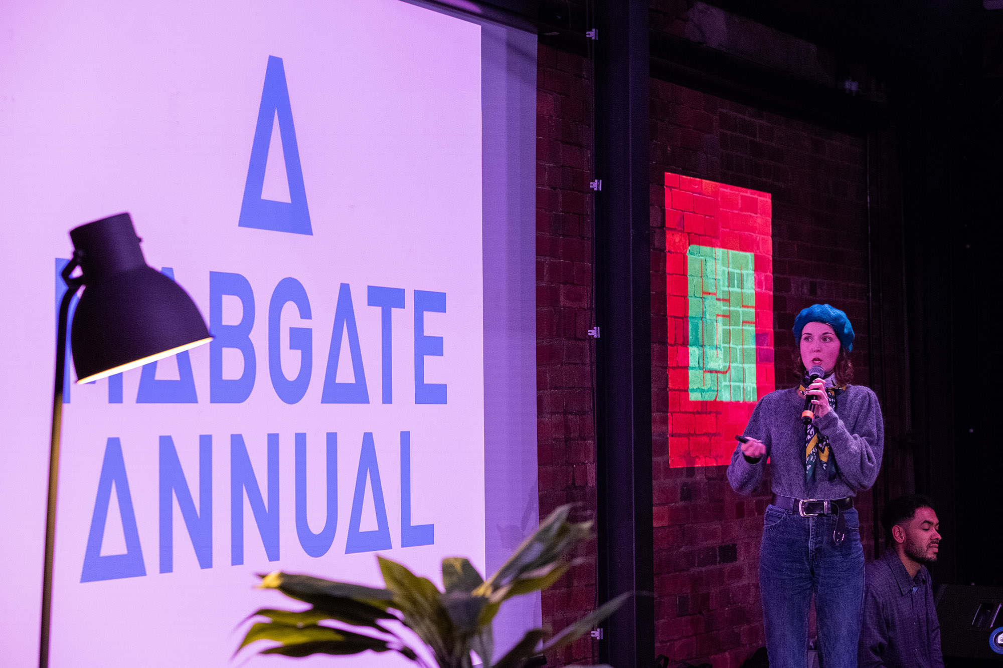 A woman stands to the right of the stage as she speaks in front of a projected presentation.