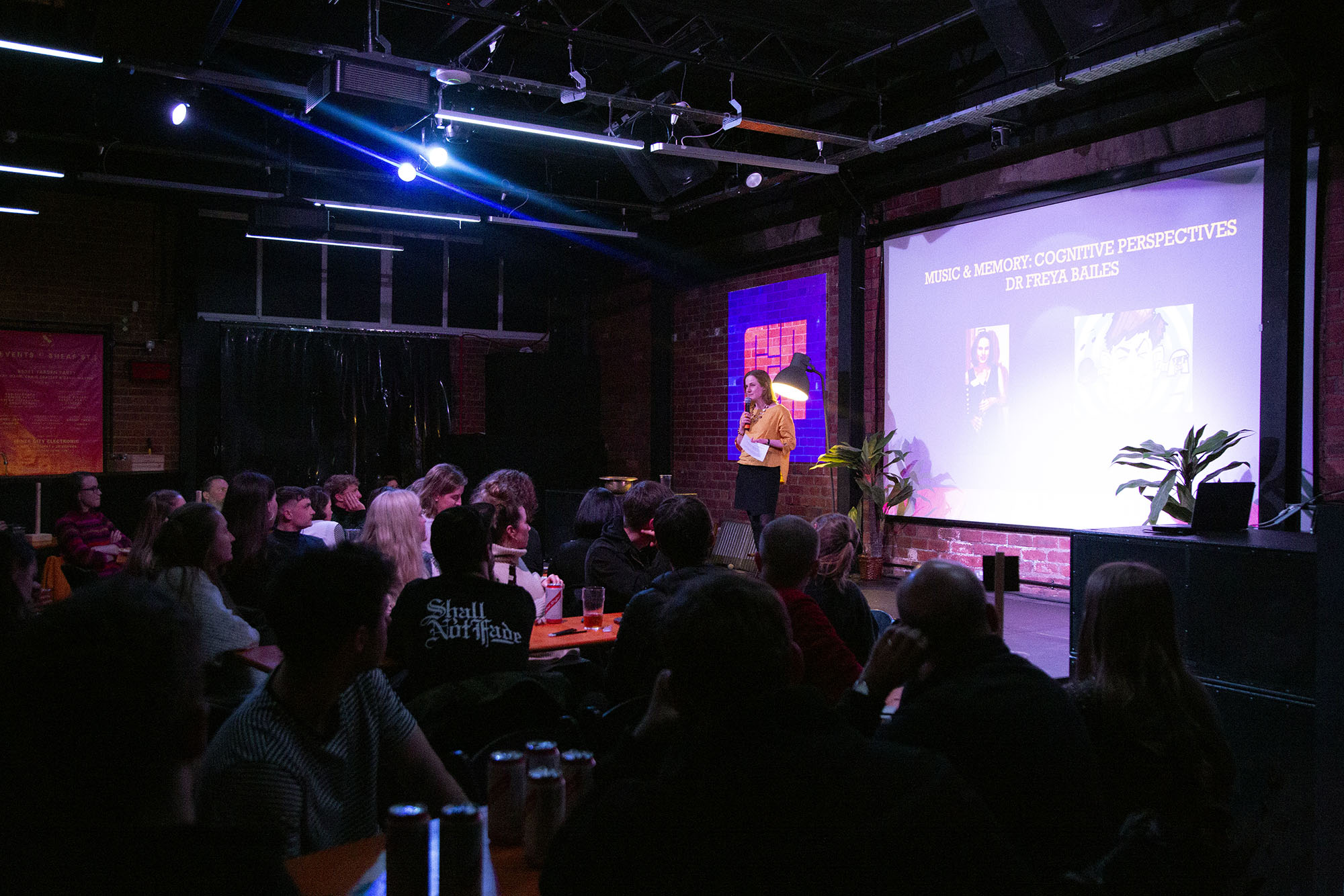 Audience watches a woman presenting in front of a slide, with the Chew the Fat logo projected on a brick wall in the background.
