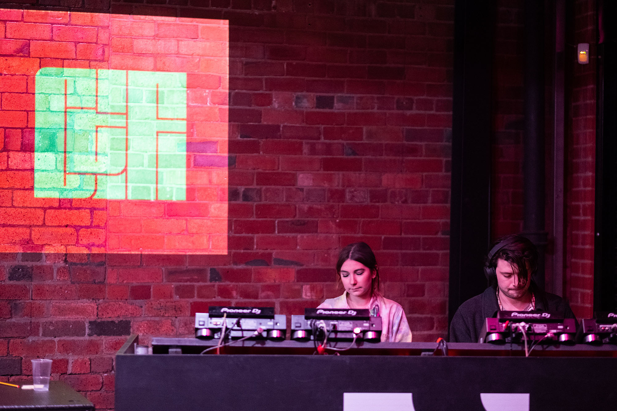 Two people playing music at a DJ booth, standing in front of a brick wall with the Chew the Fat logo projected on it.