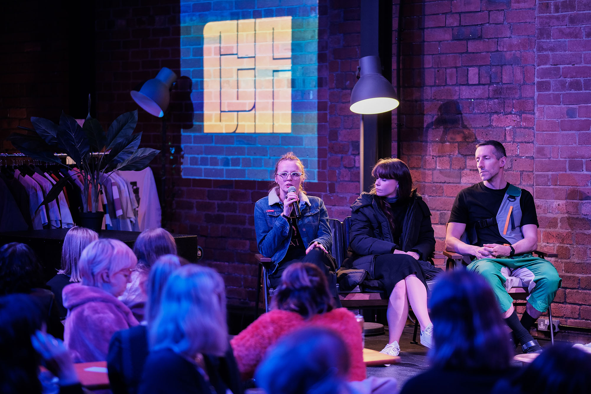Audience watches three people speaking on stage, with the Chew the Fat logo projected on a brick wall in the background.
