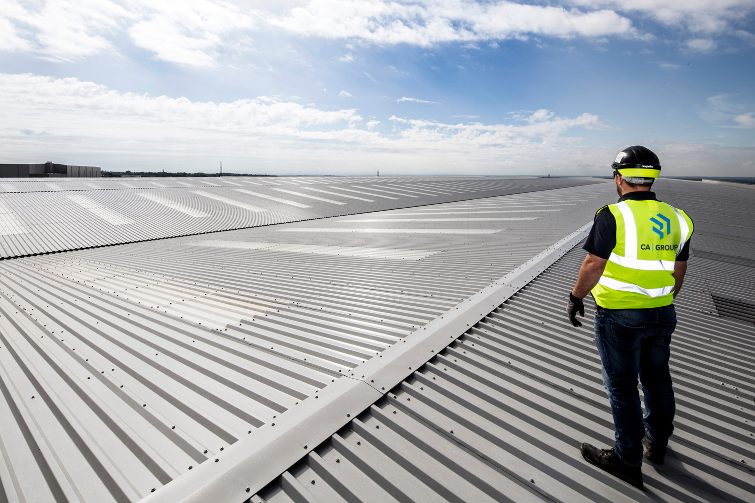 Man wearing a high-visibility jacket with the CA Group logo, with his back to the camera, looking out at a blue cloudy sky.