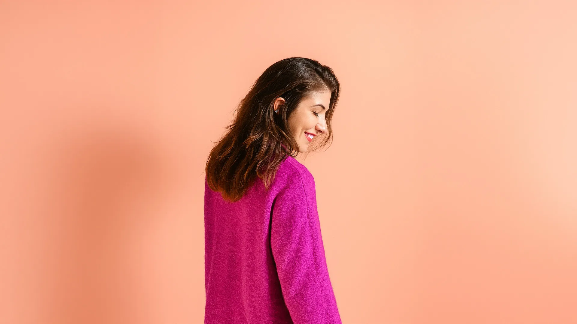 A women wearing a purple jumper with her back to the camera looking over her right shoulder smiling on a peach coloured backdrop.
