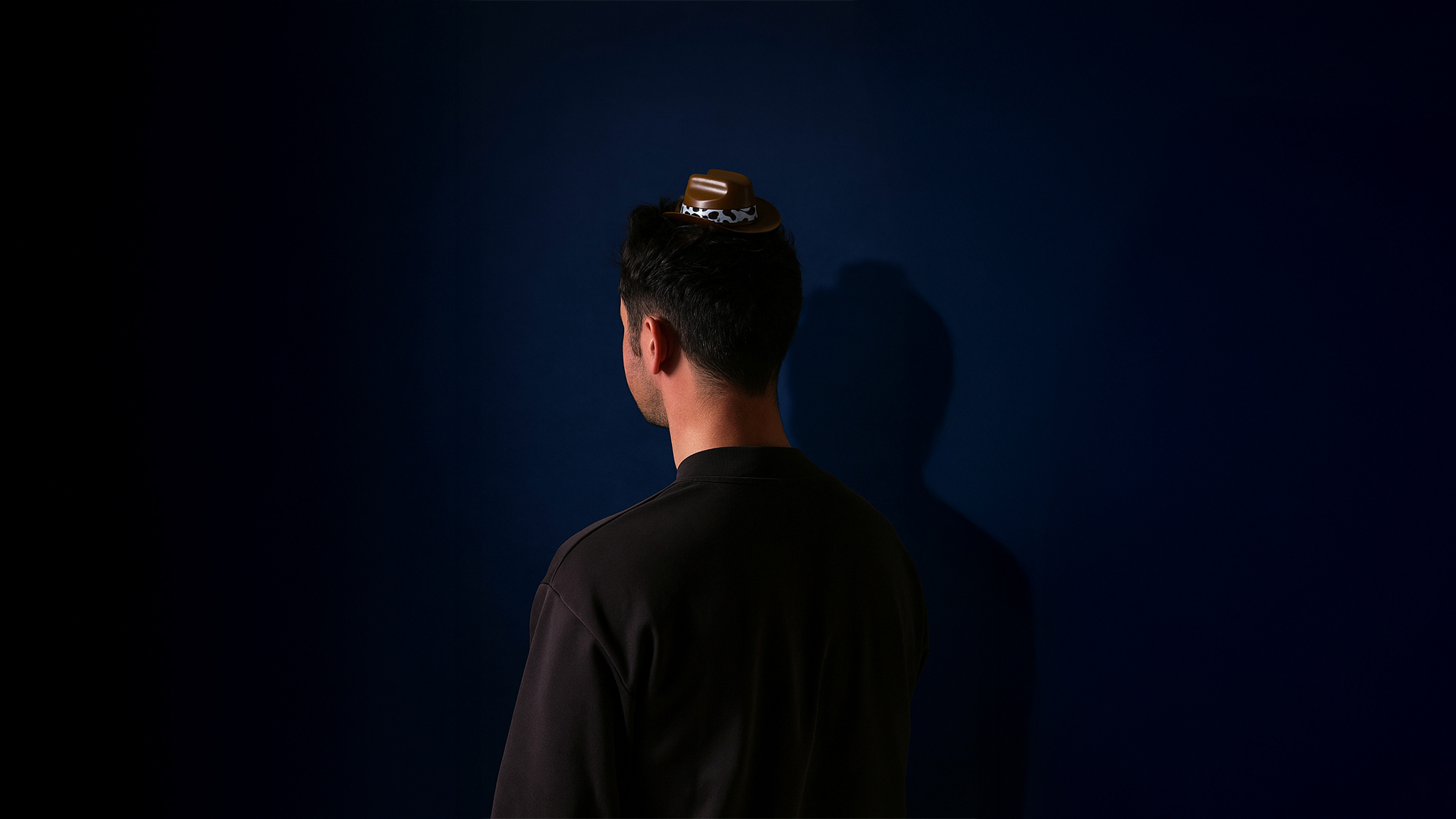 A man wearing a black t-shirt and mini brown cowboy hat with his back to the camera on a dark navy backdrop.