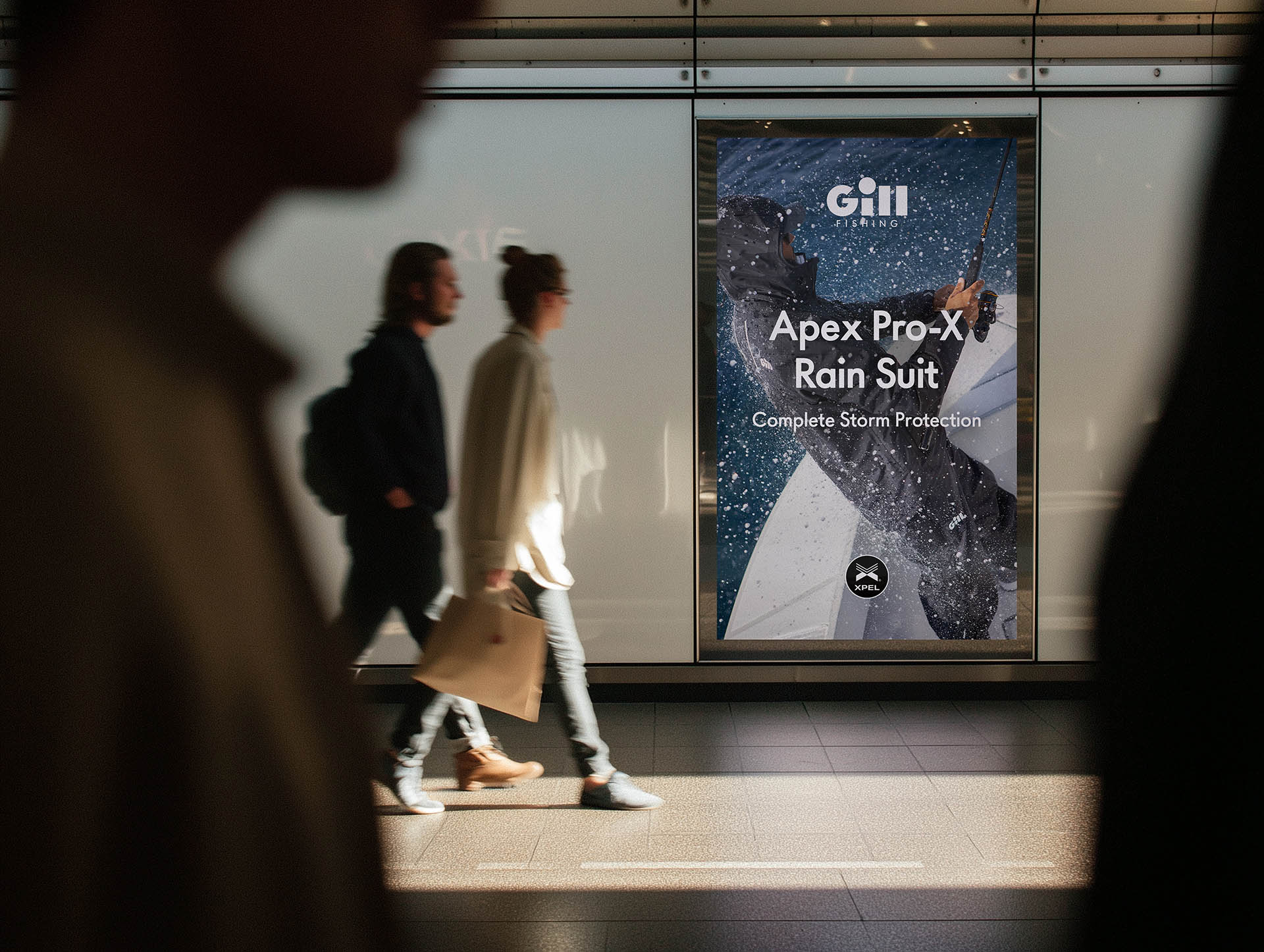 Two people walk through a subway past a digital display advertising waterproof outdoor clothing.