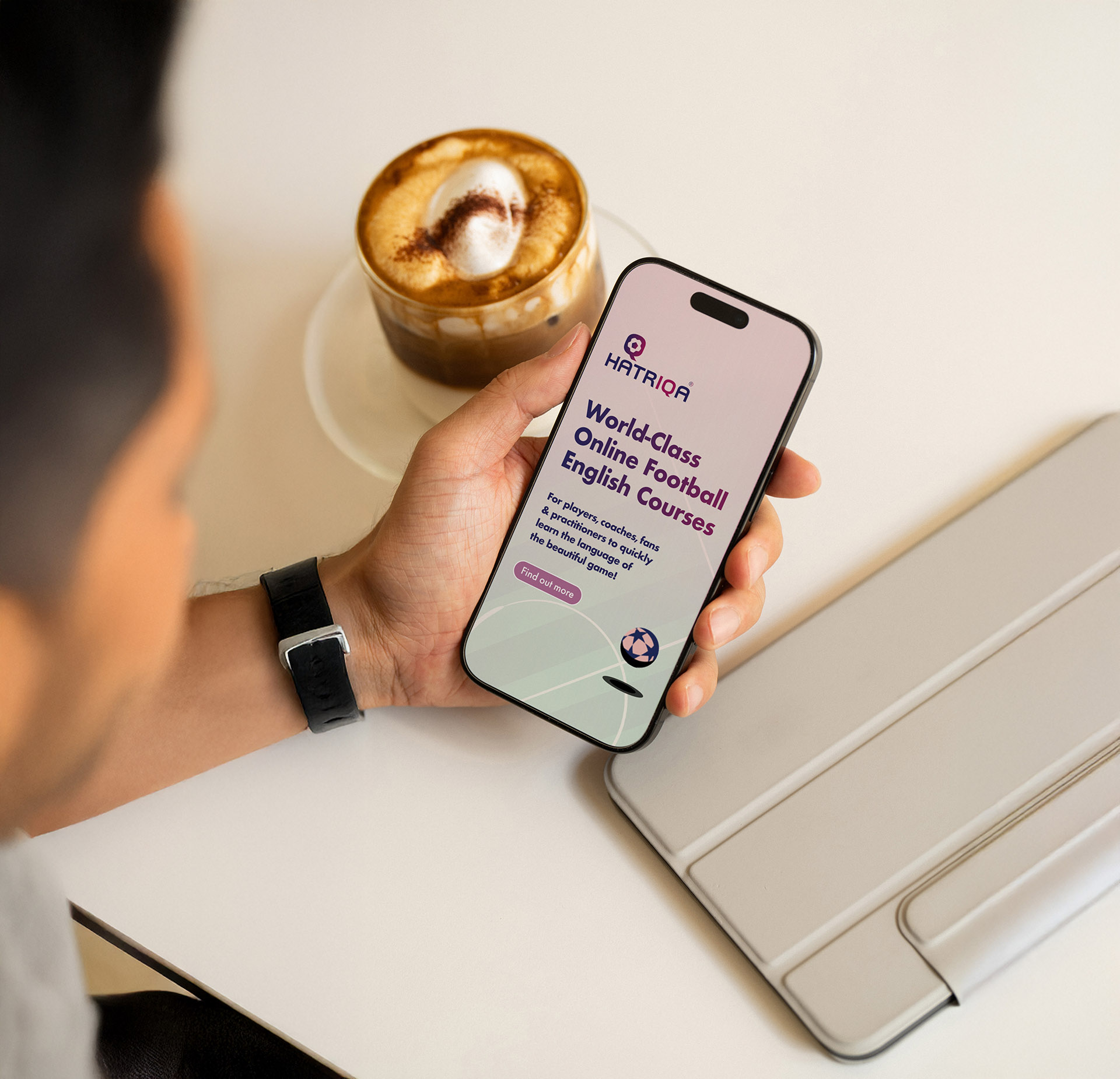Close-up of a man’s hand holding a phone, with a coffee in the background; the phone screen shows a digital advert for online English football courses.